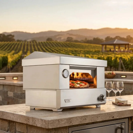 Videl Counter Top Pizza Oven, stainless steel sits on a stone patio with a vineyard and hills in the background at sunset. A pizza is baking inside, with flames visible through the oven door, and two wine glasses and a small food item are placed beside it.