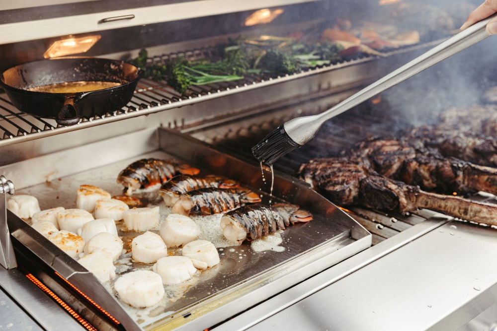Lobster tails and scallops cooking on a griddle beside steaks in an American Made Grills Muscle grill